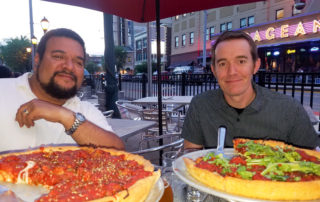 REspace Co-Founders Ryan and Eric seated at an outdoor wire table at the restaurant Pi in the Delmar Loop St. Louis, MO. 2 deep dish pizzas on stands in the foreground, the Pageant night club in the background. Photo is taken looking west at dusk.