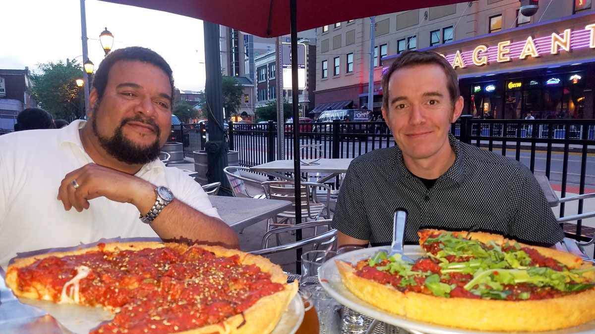 REspace Co-Founders Ryan and Eric seated at an outdoor wire table at the restaurant Pi in the Delmar Loop St. Louis, MO. 2 deep dish pizzas on stands in the foreground, the Pageant night club in the background. Photo is taken looking west at dusk.