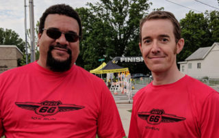 Eric and Ryan, REspace Co-founders in their red route 66 10k volunteer shirts. Photo is full frame with small amounts of the race finish line in the background visible.