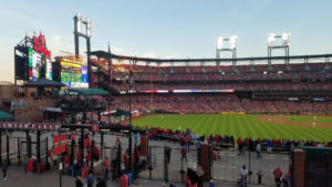 A view of the Busch stadium from the balcony at the Ball Park Village. The centerfield gate, scoreboard, outfield, and pitchers mound along with the bleachers along the first base line are visible. The lights are already on under a pale blue sky as the sun begins to set outside of the photo on the far right.