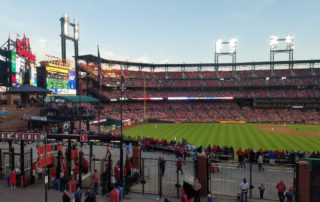 A view of the Busch stadium from the balcony at the Ball Park Village. The centerfield gate, scoreboard, outfield, and pitchers mound along with the bleachers along the first base line are visible. The lights are already on under a pale blue sky as the sun begins to set outside of the photo on the far right.
