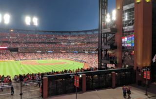 A view of the Busch stadium from the balcony at the Ball Park Village. The view is looking at home plate; the entire infield is visible along with the bleachers along the first base line wrapping around to the 3rd base foul post. The sky is a deep blue from the last rays of light from the already set sun.